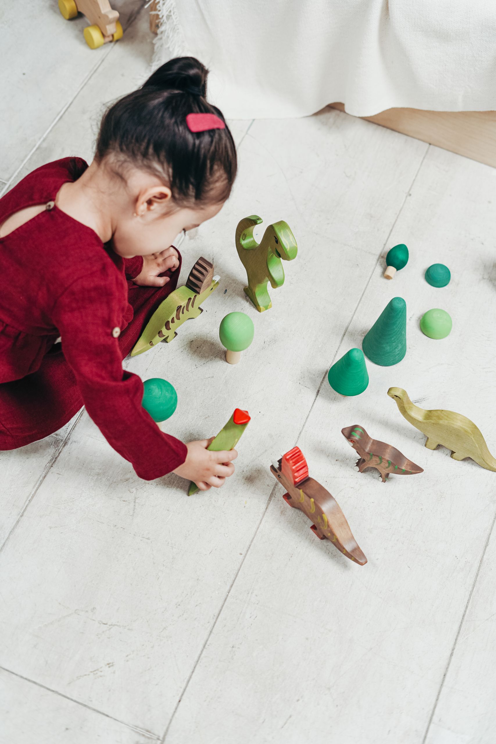 A child in red dress playing with wooden dinosaur toys indoors, sparking creativity and imagination.